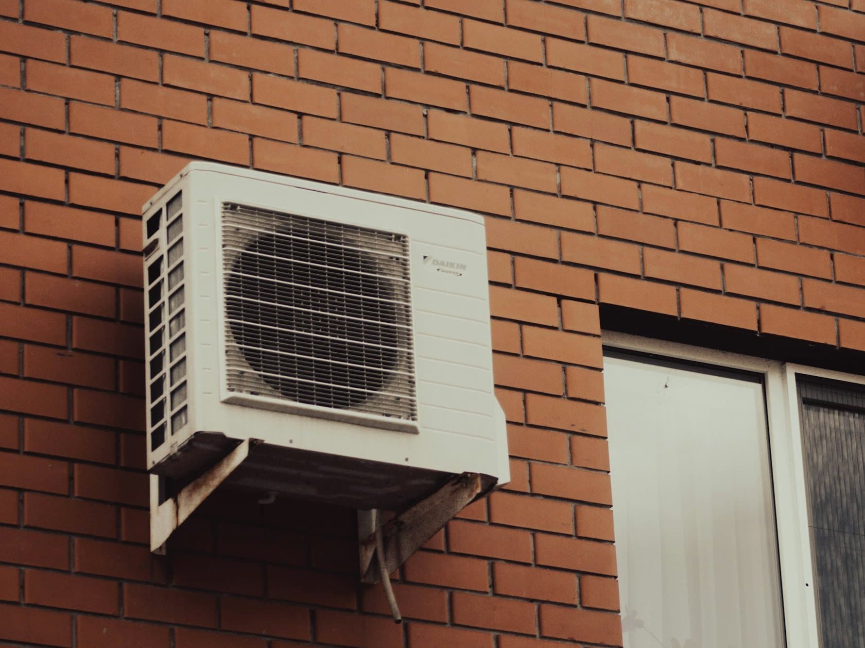 The outdoor unit of a split system heat pump, mounted on a bracket on a red-brick wall beside a window.