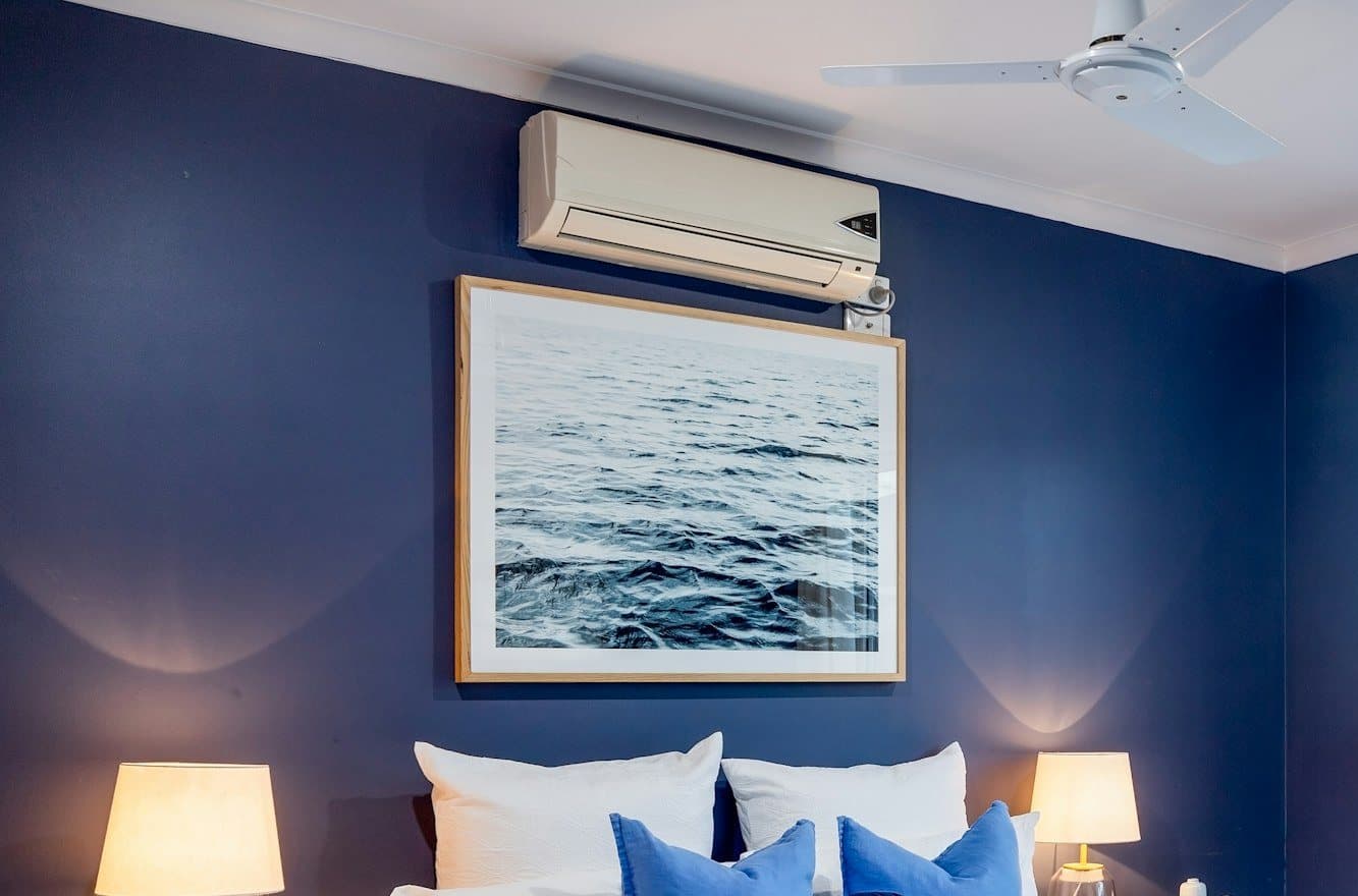A white wall-mounted indoor unit high on a bedroom wall, above a framed picture and the bed.